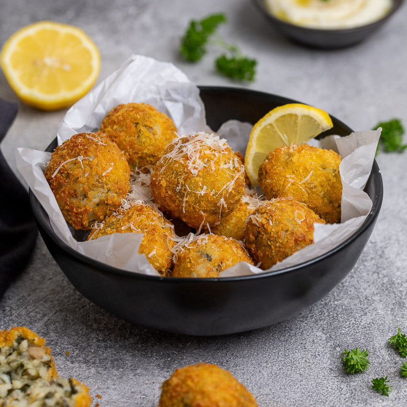 A bowl of golden-brown arancini (Italian rice balls) garnished with grated cheese and lemon slices, with herbs scattered around on a grey background.