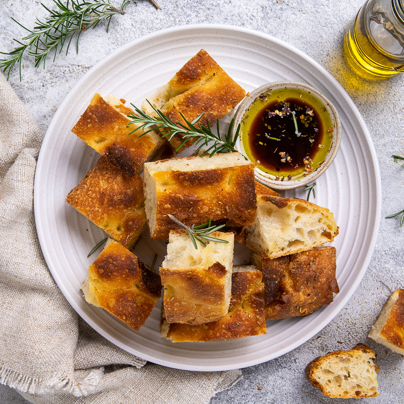 Platter of focaccia bread slices with a small bowl of olive oil and rosemary on a light background