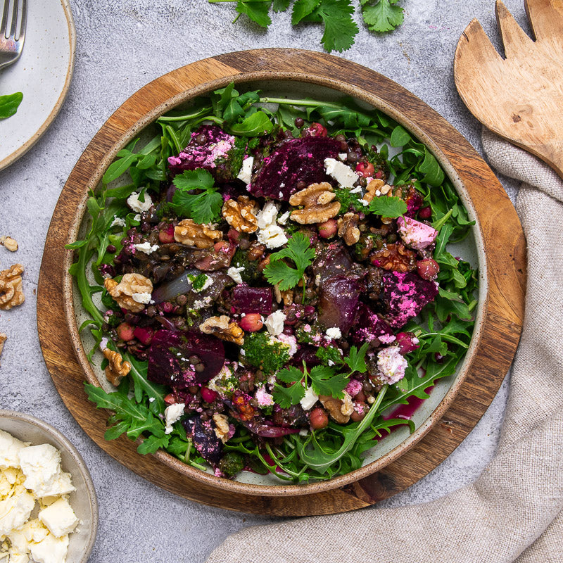 A prepared dish of French lentils, roast beetroots, caramelised red onion, walnuts, and marinaded feta, served on a bed of greens in a wooden bowl.