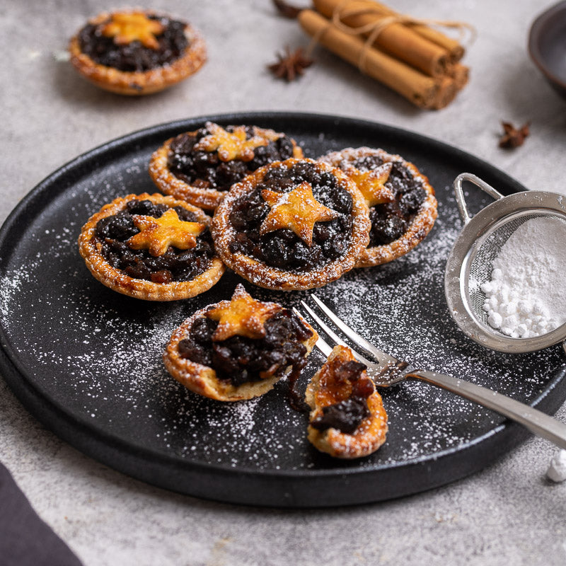Four fruit mince pies on a black plate, topped with icing sugar and cinnamon, with a spoon with icing sugar on the side.