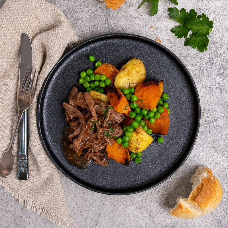 Plated dish of roast beef, sweet potatoes, and green peas on a gray plate with a knife and fork.