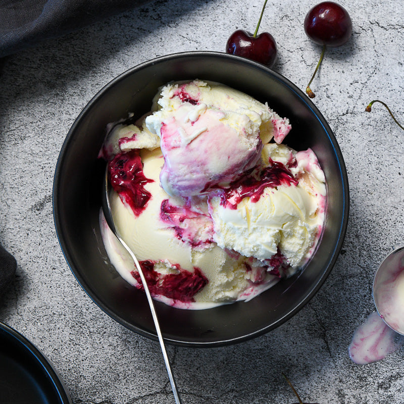 A bowl of Yogurt Sour Cherry Gelato with a spoon and cherries on a gray background