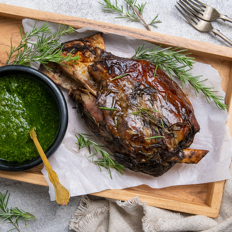 A plate with a cooked lamb shoulder piece, served with a side of green salsa verde and garnished with rosemary.
