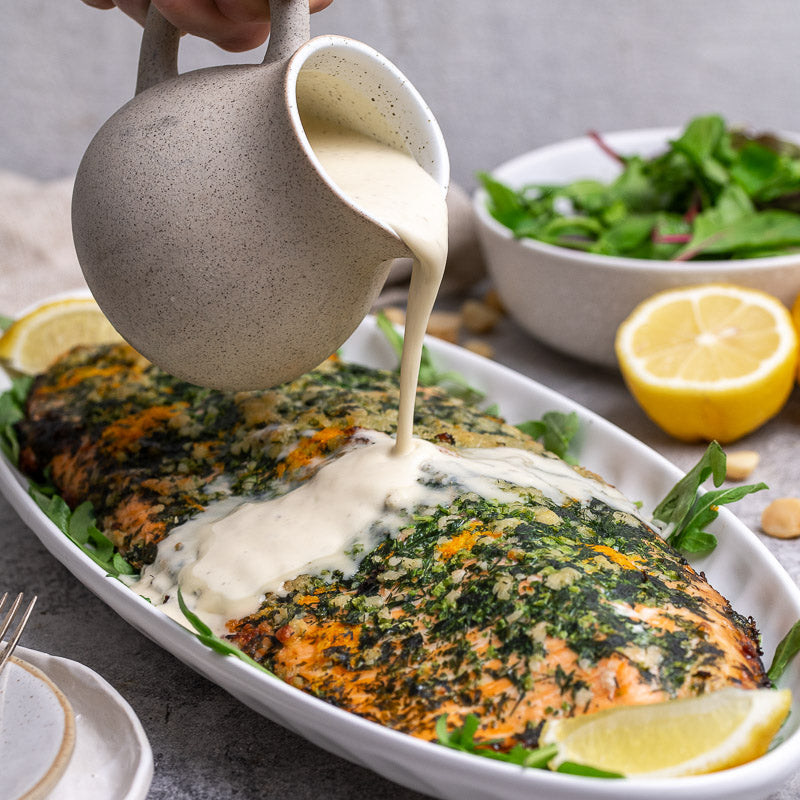 A salmon fillet with macadamia crust on a bed of greens, being drizzled with lemon myrtle velouté sauce.