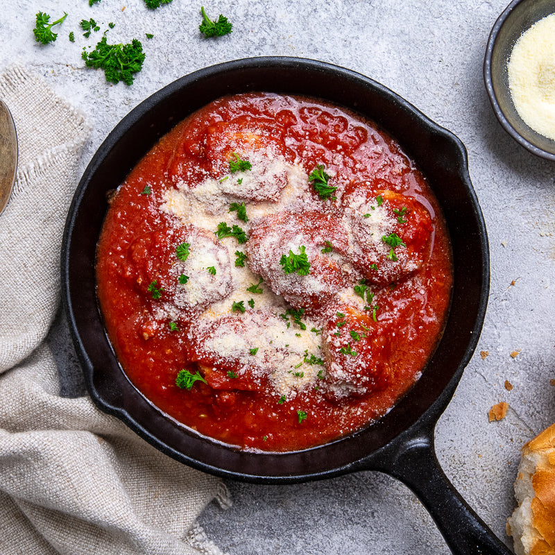 Skillet with meatballs in tomato sauce on a gray surface with herbs and bread.