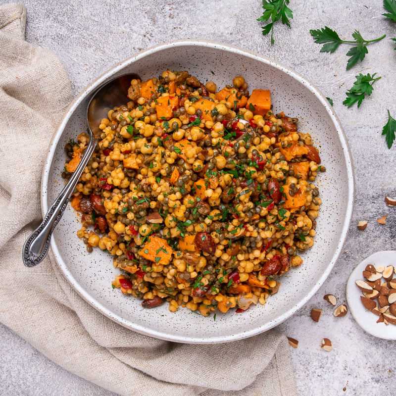 A bowl of cooked couscous salad with sweet potato, lentils, herbs, and dried fruit, served on a light stone surface with a spoon and small garnish props.