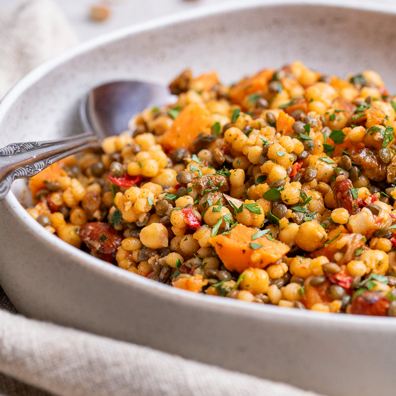 A close-up of a cooked lentil and vegetable salad in a light gray ceramic bowl with a spoon, featuring yellow lentils, orange butternut squash, green herbs, and red peppers on a neutral tabletop.