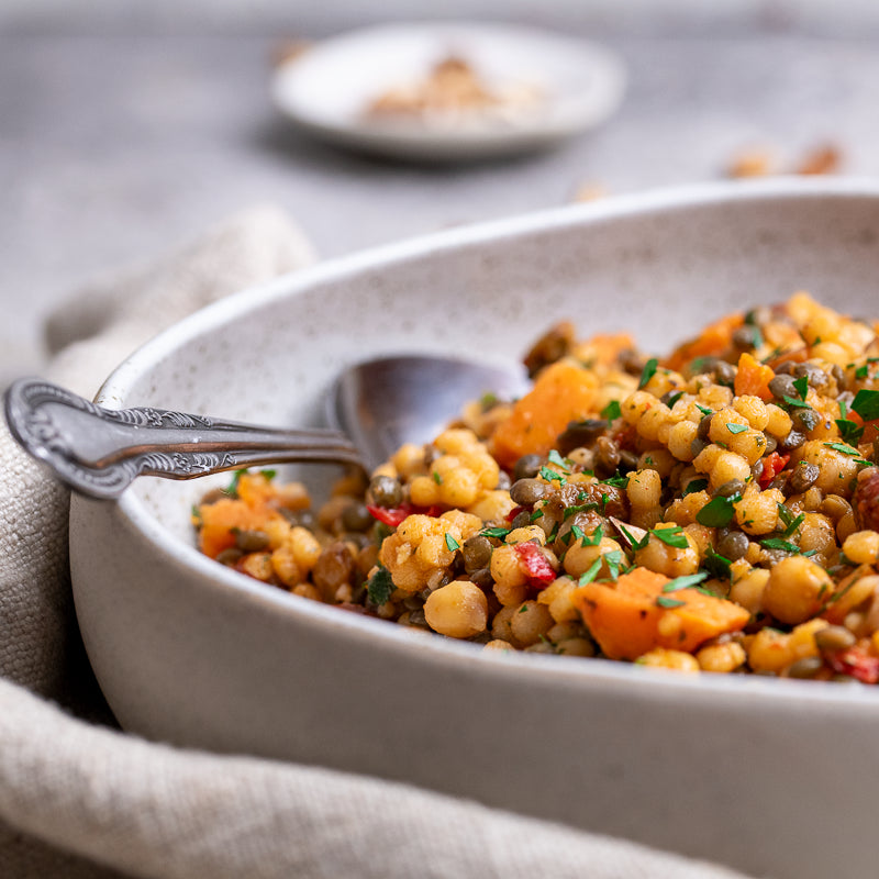 A close-up of a savory chickpea and sweet potato salad served in a speckled off-white ceramic bowl with a silver spoon, set on a textured beige cloth over a gray surface.