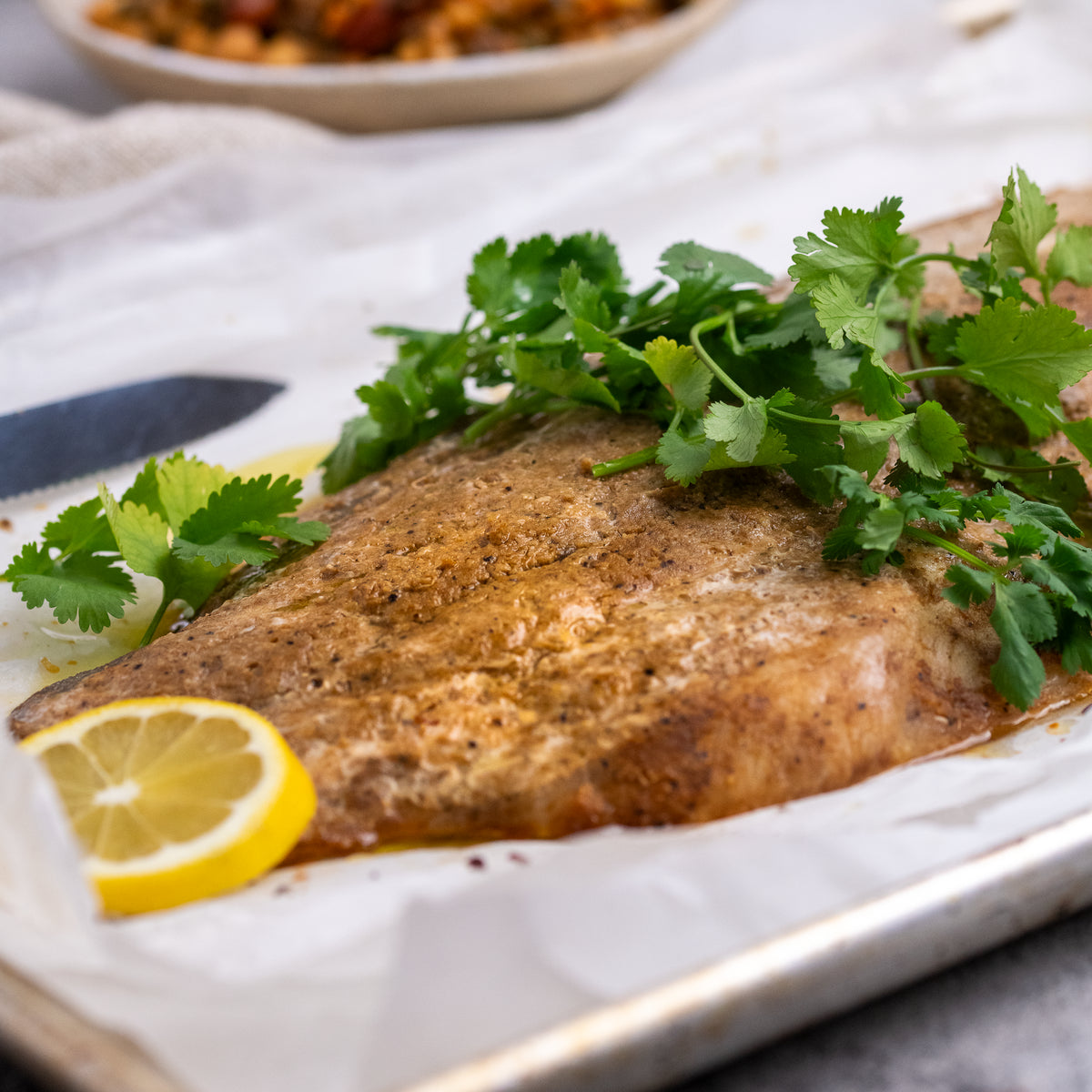 A cooked fish fillet on a baking sheet with cilantro and a lemon slice, photographed in a real kitchen setting with a blurred bowl in the background.