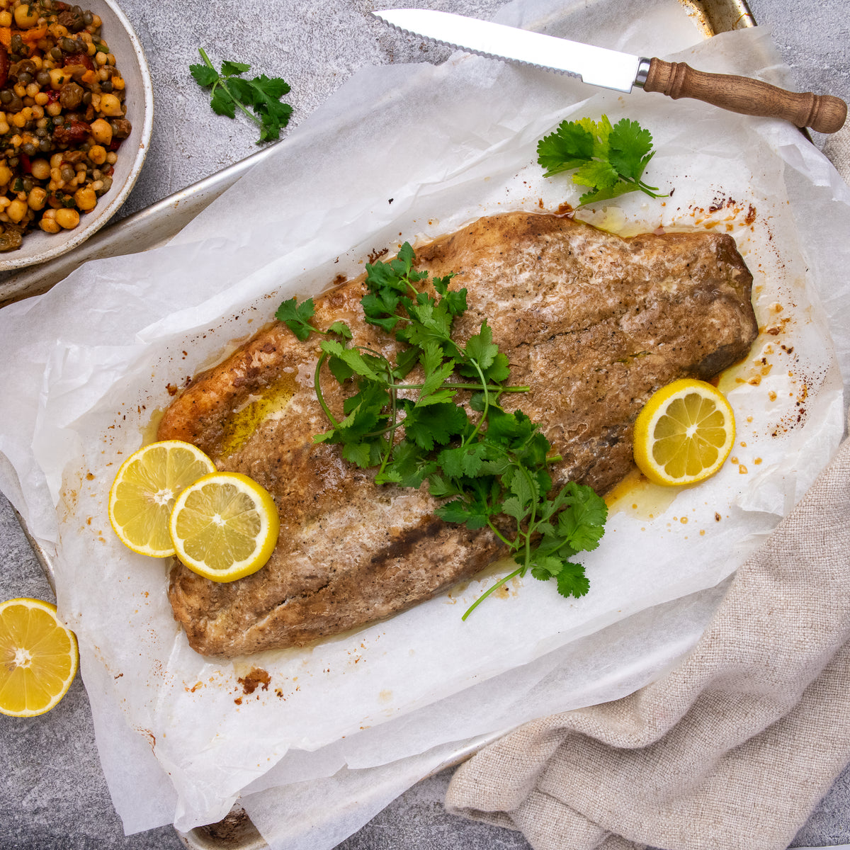 A cooked fish fillet on parchment paper, garnished with fresh cilantro and lemon slices, styled with a knife, a bowl of chickpeas, and a beige cloth on a gray surface.