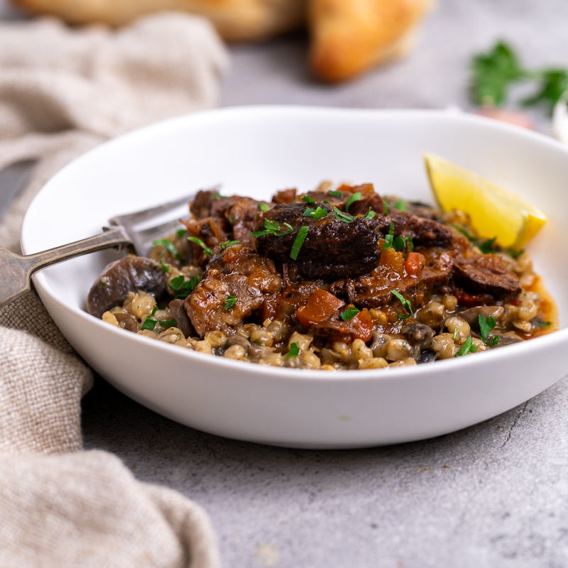 White bowl of beef cheeks and orzoni risotto with lemon wedges on a gray surface.