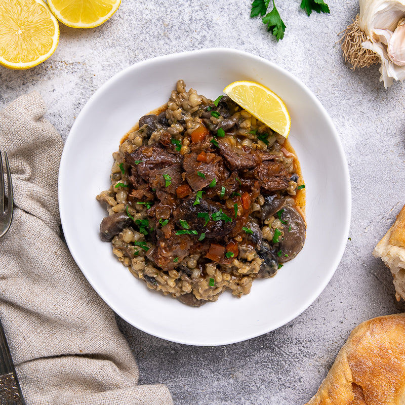 White bowl of beef cheeks and orzoni risotto with lemon wedges on a gray surface.