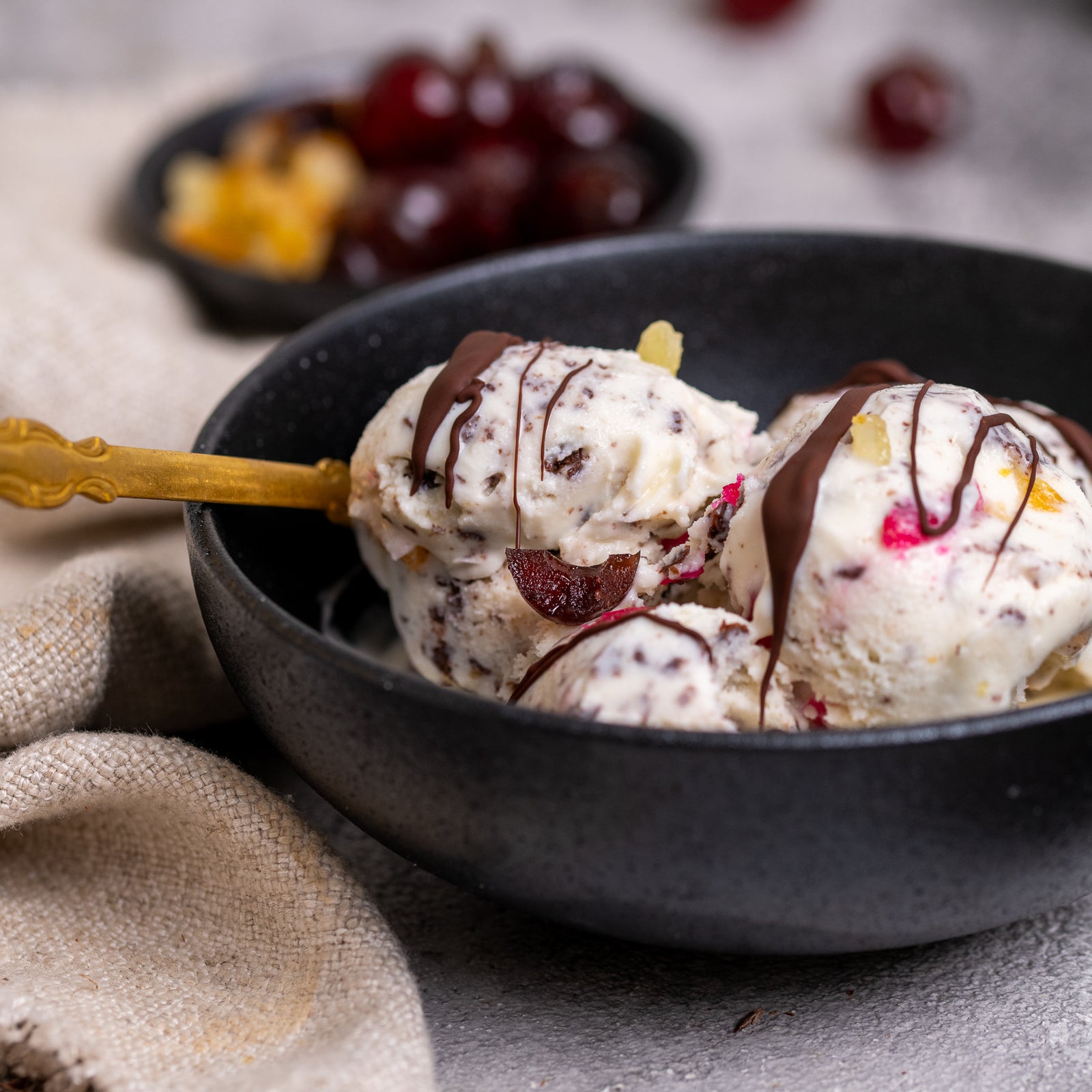 Bowl of sicilian cassata ice cream with fruits and nuts on a textured surface with a gold spoon and chocolate bar.