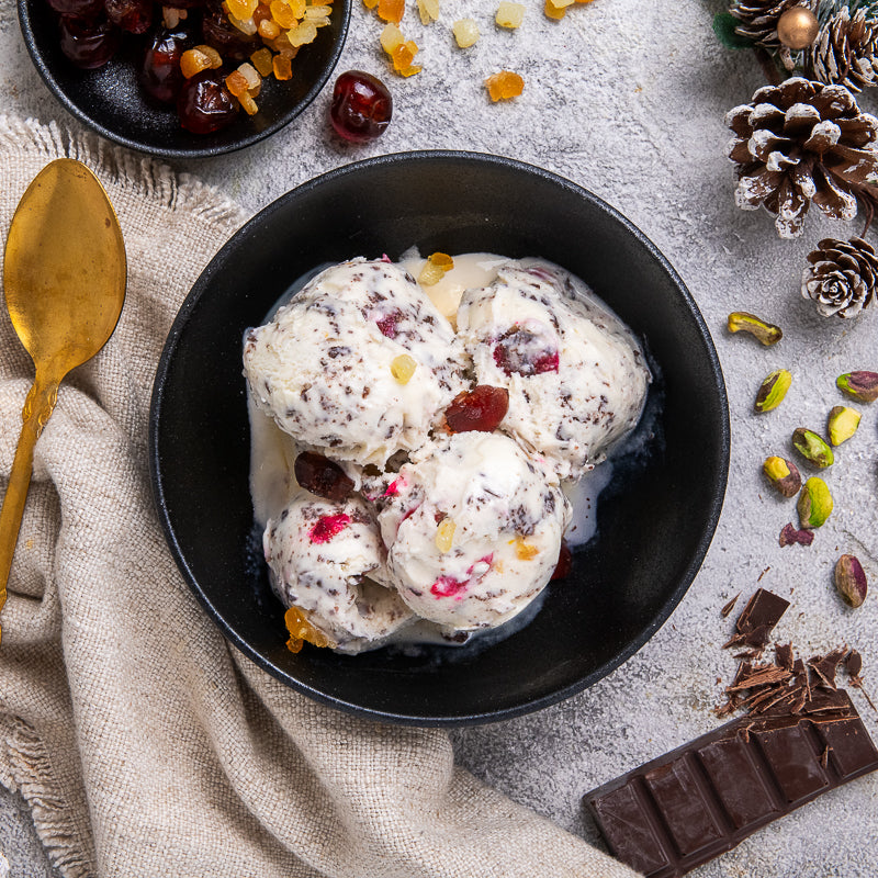 Bowl of sicilian cassata ice cream with fruits and nuts on a textured surface with a gold spoon and chocolate bar.