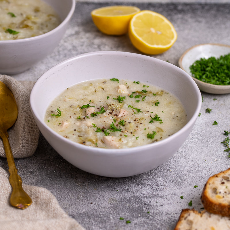 White bowl of chicken and rice soup with lemon and bread on a gray surface