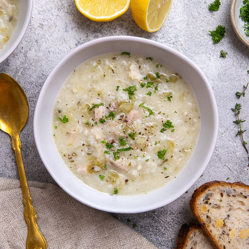 White bowl of chicken and rice soup with lemon and bread on a gray surface