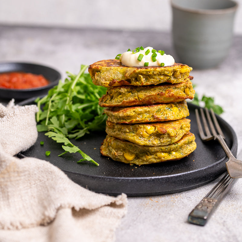 Stack of corn fritters on a black plate with arugula and sour cream garnish.