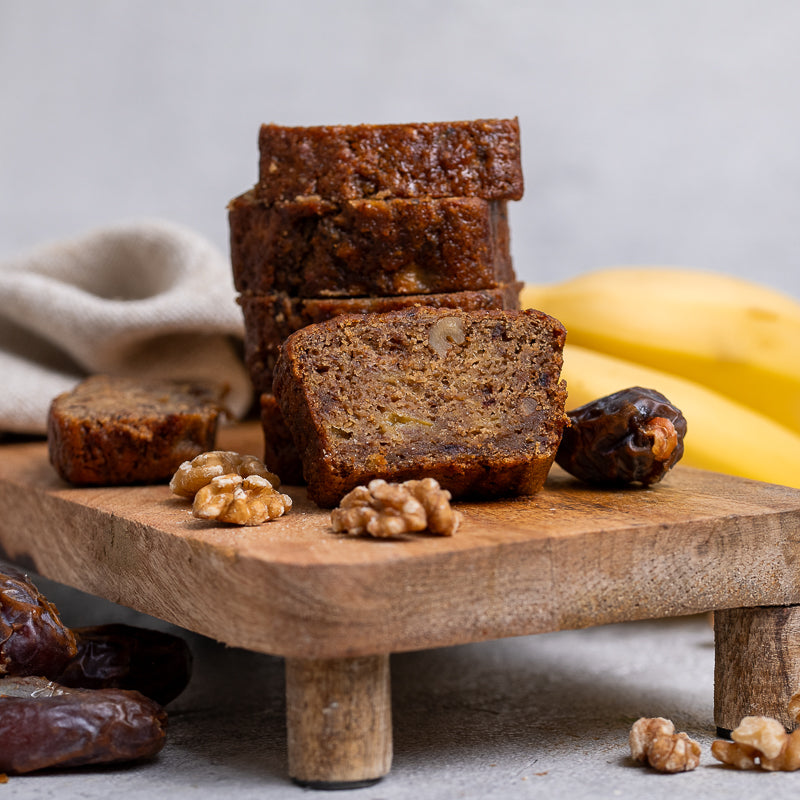 Sliced banana bread on a wooden board with bananas and dates in the background