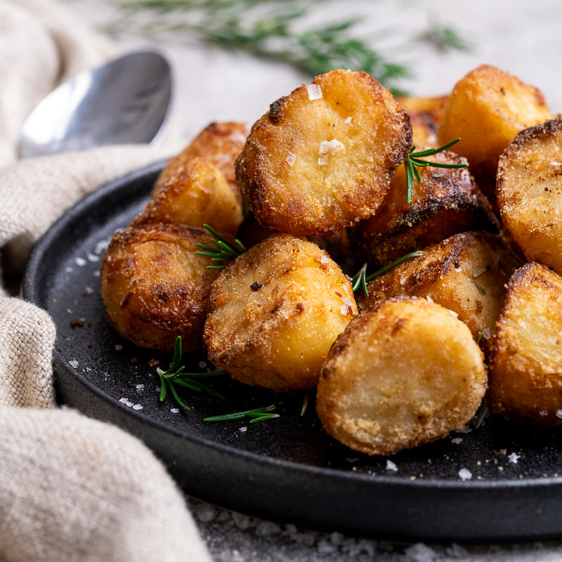 Crispy golden-brown roasted potato chunks on a black plate, garnished with fresh rosemary sprigs and coarse sea salt, with a beige cloth napkin and spoon in the background.