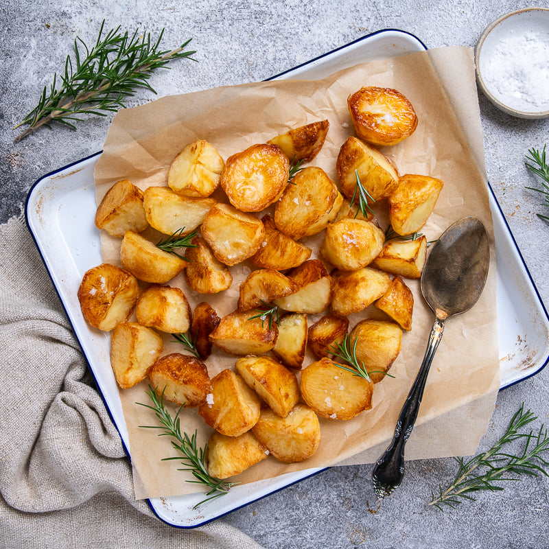 Roasted potatoes cooked in duck fat on a tray with rosemary and a spoon, on a light gray surface.