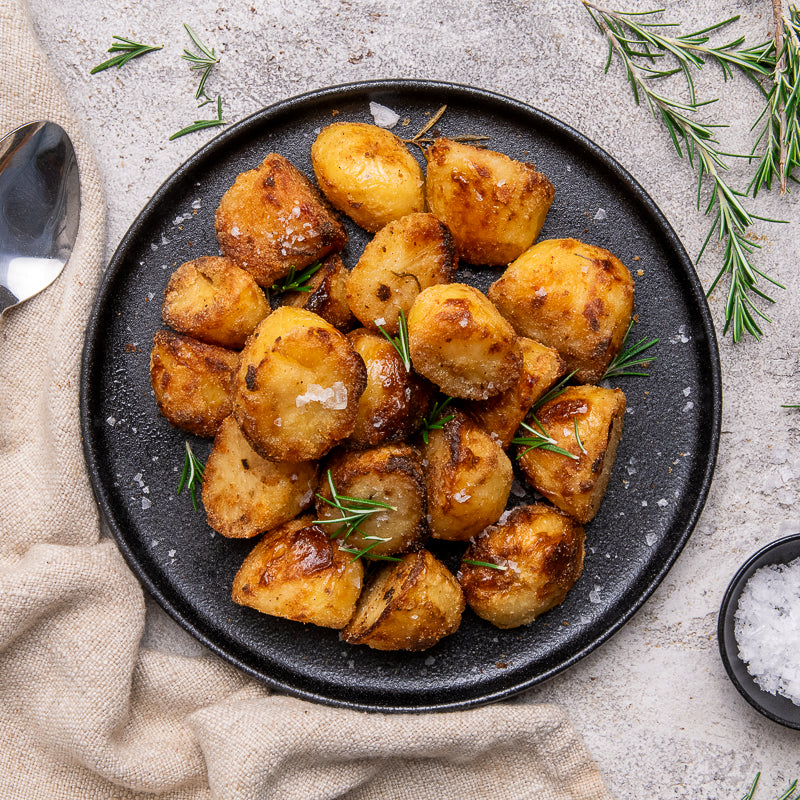 Roasted baby potatoes with crispy browned edges, garnished with fresh rosemary sprigs and coarse sea salt, served on a dark gray speckled plate. The plate is set on a textured light gray stone surface with a beige linen napkin, a spoon on the left, and a small bowl of salt on the bottom right.