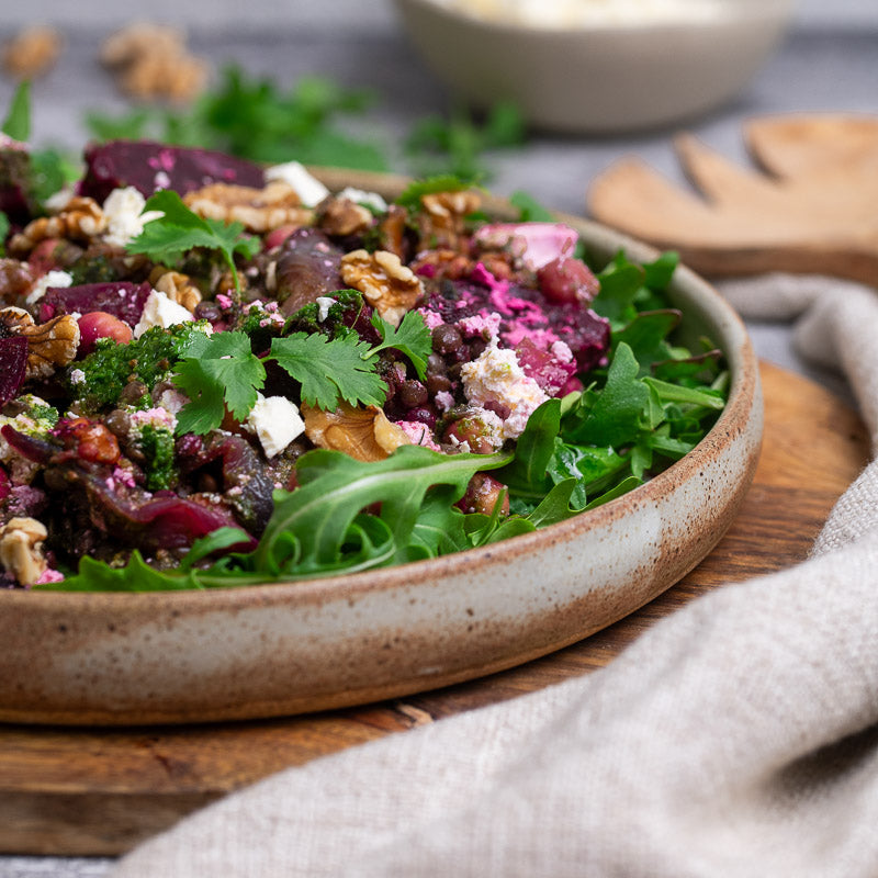 A prepared dish of French lentils, roast beetroots, caramelised red onion, walnuts, and marinaded feta, served on a bed of greens in a wooden bowl.