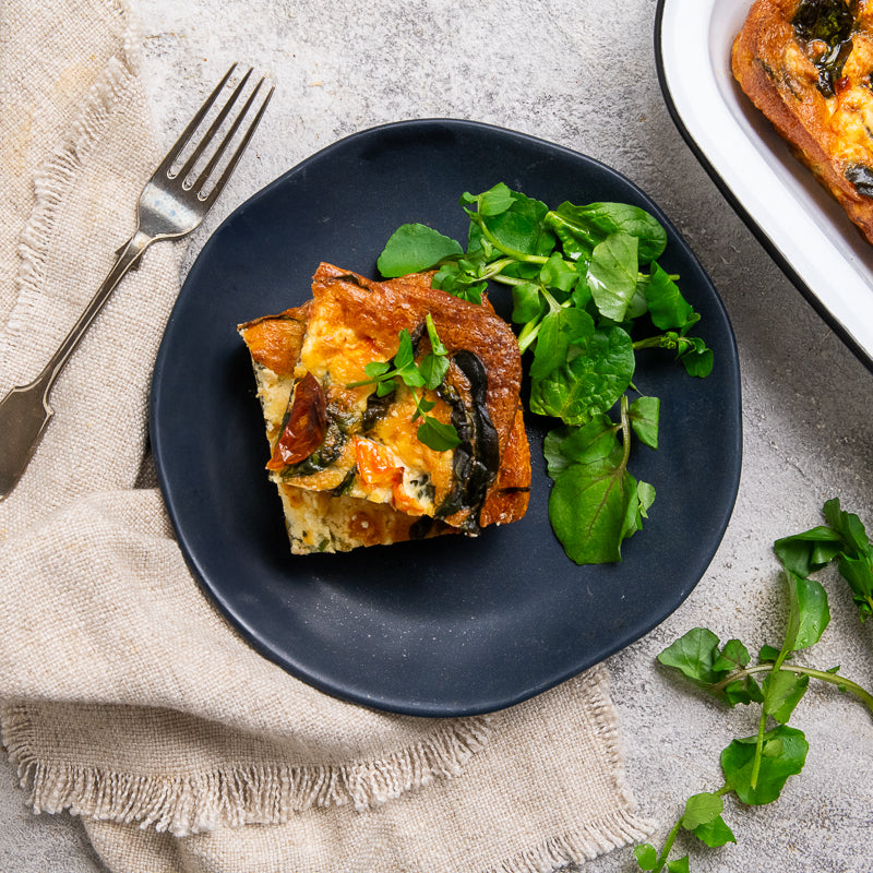 Square of vegetable lasagna on a dark plate with a side of greens, on a textured surface.
