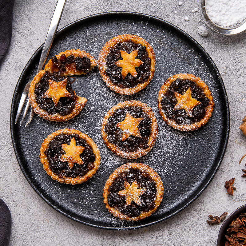 Four fruit mince pies on a black plate, topped with icing sugar and cinnamon, with a spoon with icing sugar on the side.