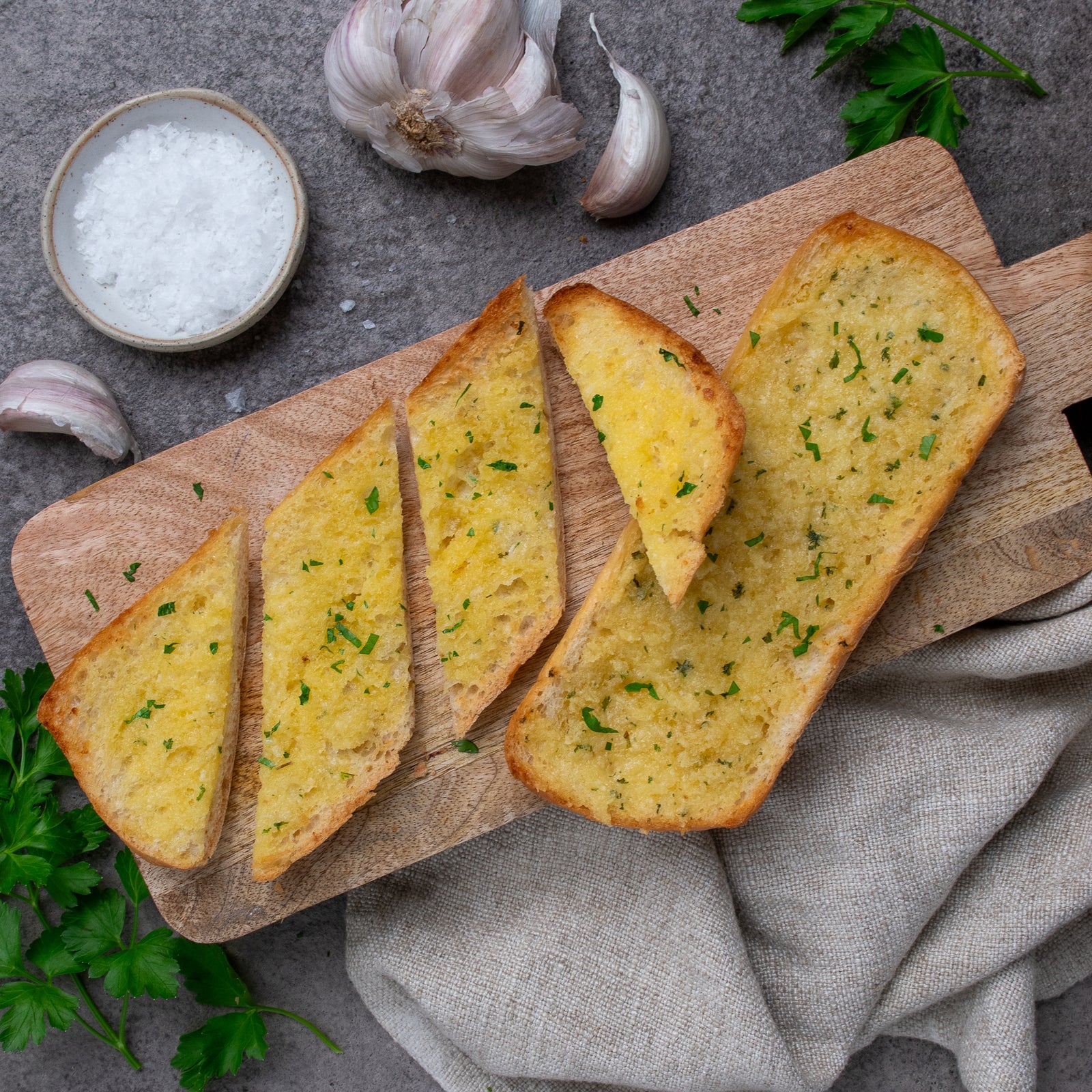 Nat's Garlic Ciabatta Bread on a rustic chopping board, sliced with parsely and garlic in the backround