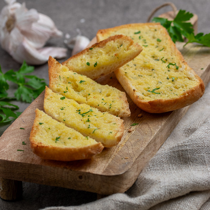 Nat&#39;s Garlic Ciabatta Bread on a rustic chopping board, sliced with parsely and garlic in the backround