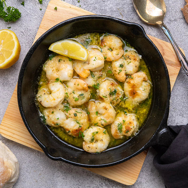 Skillet of prawns in a garlic butter sauce with lemon wedges on a wooden board