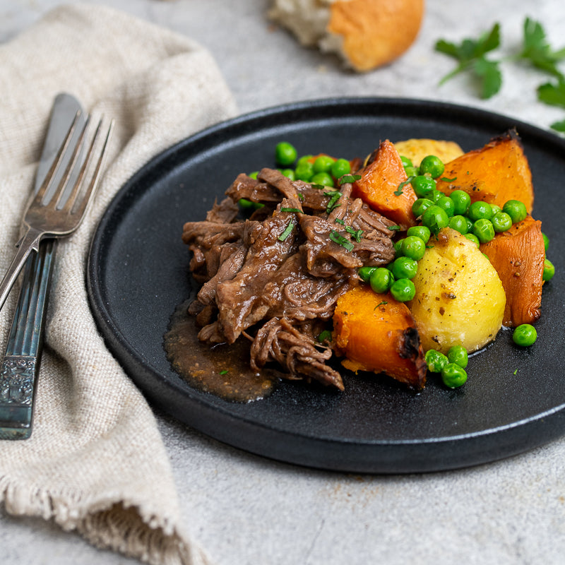 Plated dish of roast beef, sweet potatoes, and green peas on a gray plate with a knife and fork.