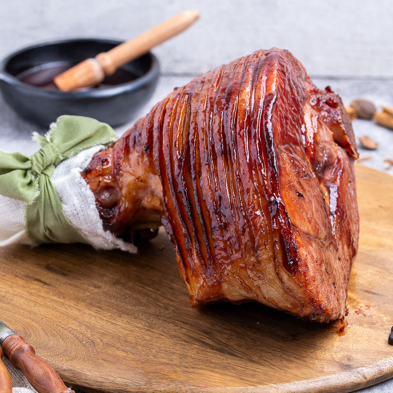 A glazed ham leg with a glossy reddish-brown bark, tied with a green cloth and white paper at the bone, resting on a wooden cutting board with a small bowl and brush in the background.