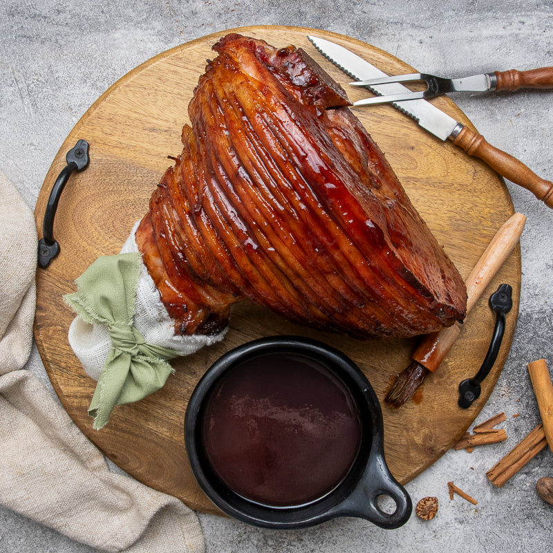 A glazed ham on a round wooden serving board with carving knives, a small bowl of sauce, and garnish on a gray stone surface.