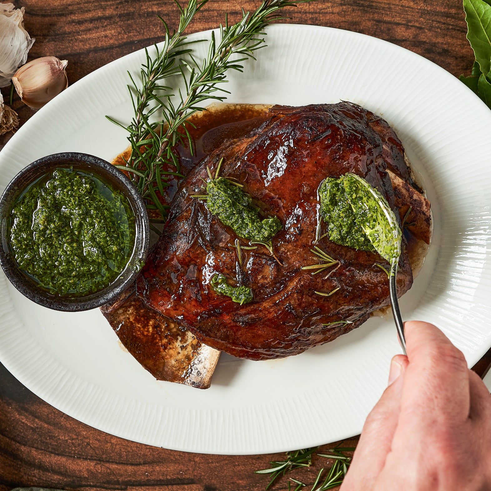 A plate with a cooked lamb shoulder piece, served with a side of green salsa verde and garnished with rosemary.