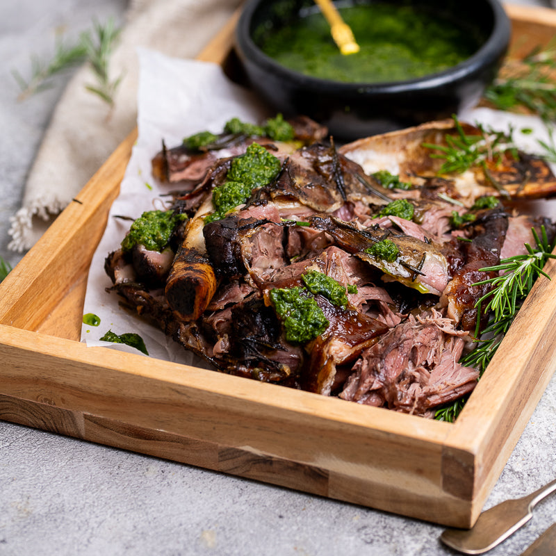 A plate with a cooked lamb shoulder piece, served with a side of green salsa verde and garnished with rosemary.