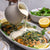 A salmon fillet with macadamia crust on a bed of greens, being drizzled with lemon myrtle velouté sauce.