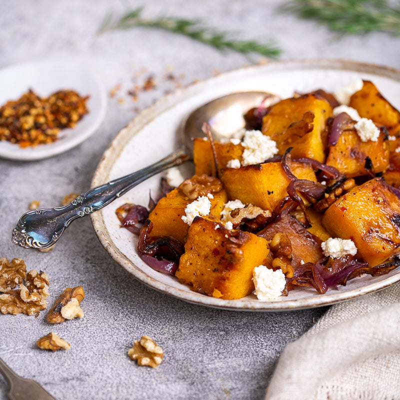Plate of roasted pumpkin cubes with maple syrup onions and feta cheese on a textured surface.