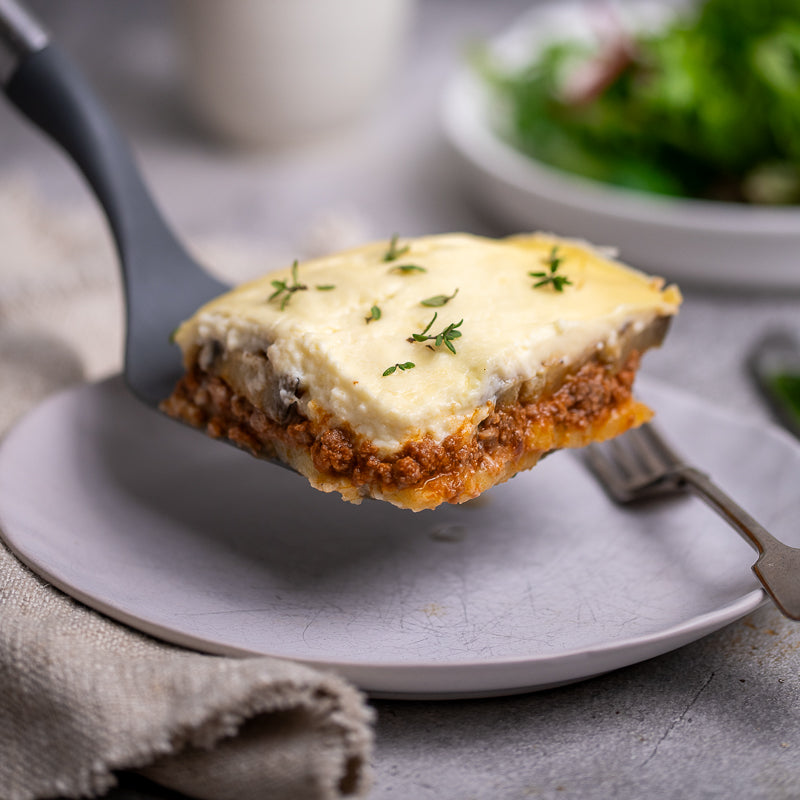 Slice of moussaka on a plate with a fork, placed on a textured surface.