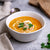 A bowl of Mulligatawny Soup topped with cream and coriander, alongside slices of bread and a spoon, on a grey background.