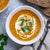 A bowl of Mulligatawny Soup topped with cream and coriander, alongside slices of bread and a spoon, on a grey background.