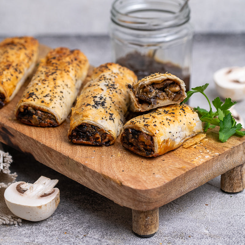 Baked mushroom rolls on a wooden board with a jar of dipping sauce and mushrooms in the background.