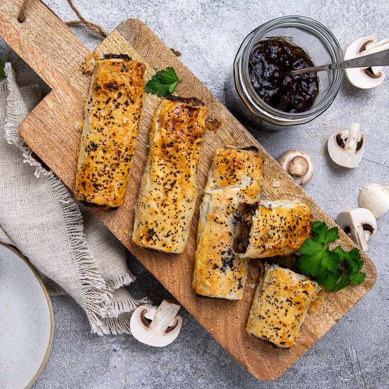 Baked mushroom rolls on a wooden board with a jar of dipping sauce and mushrooms in the background.