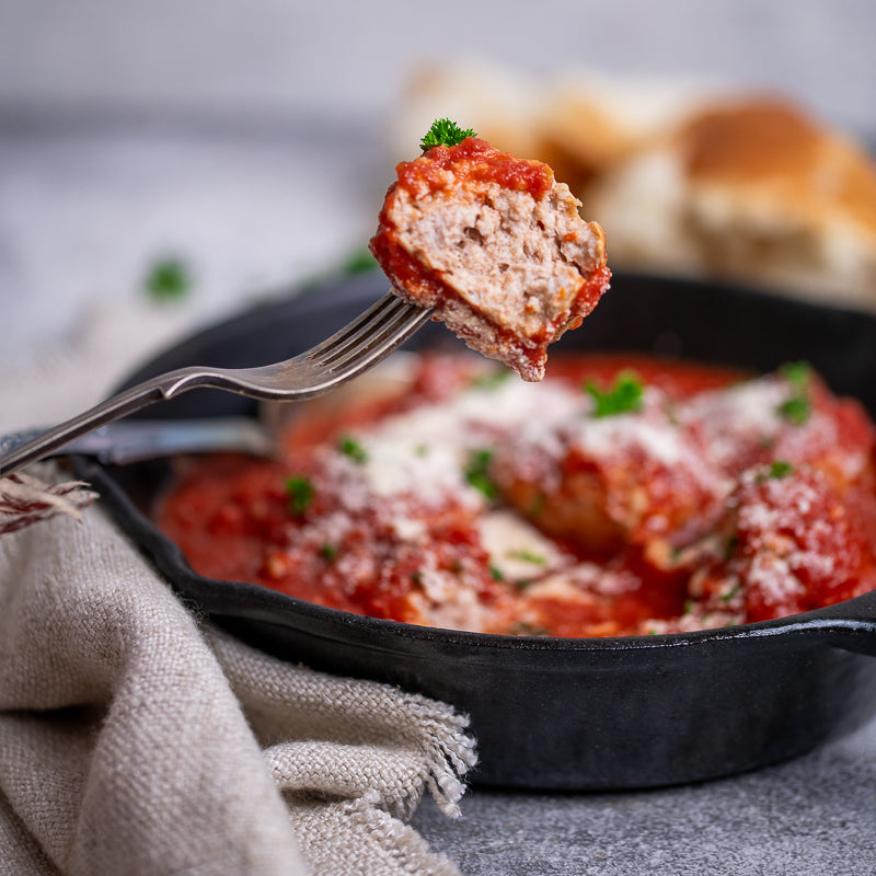 Skillet with meatballs in tomato sauce on a gray surface with herbs and bread.