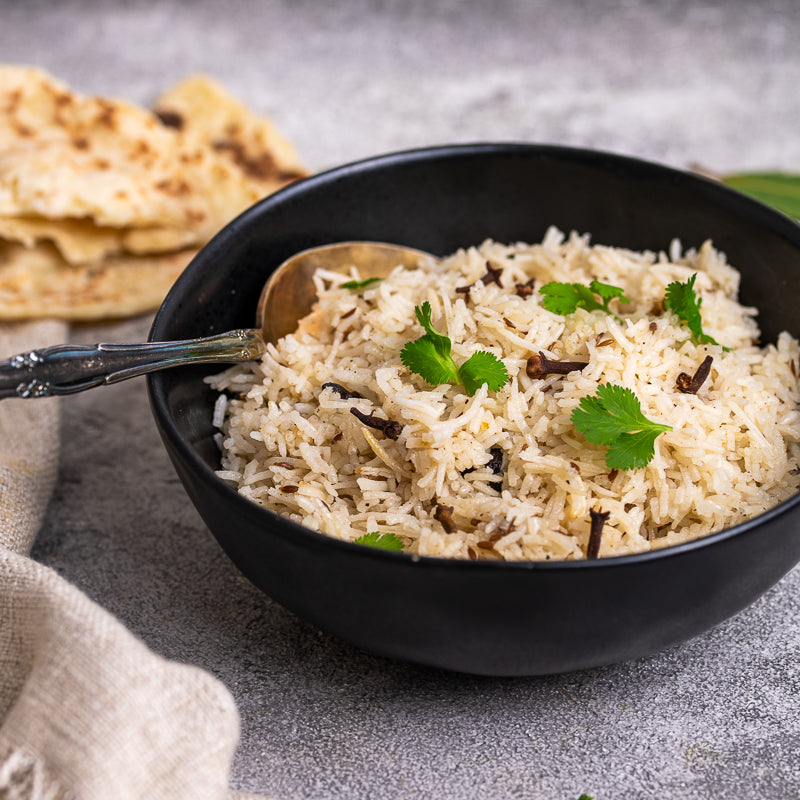 Black bowl of rice with a spoon, garnished with cilantro, on a gray surface with naan bread.