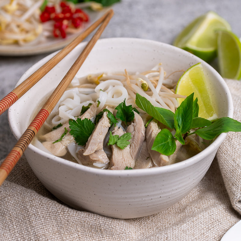 A bowl of Vietnamese chicken Pho garnished with Thai basil, bean sprouts, lime wedge, and chilli, served with hoisin and sriracha sauce on the side.