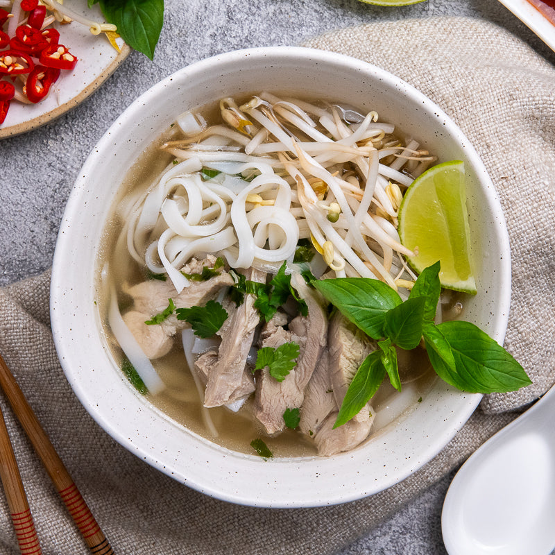 A bowl of Vietnamese chicken Pho garnished with Thai basil, bean sprouts, lime wedge, and chilli, served with hoisin and sriracha sauce on the side.