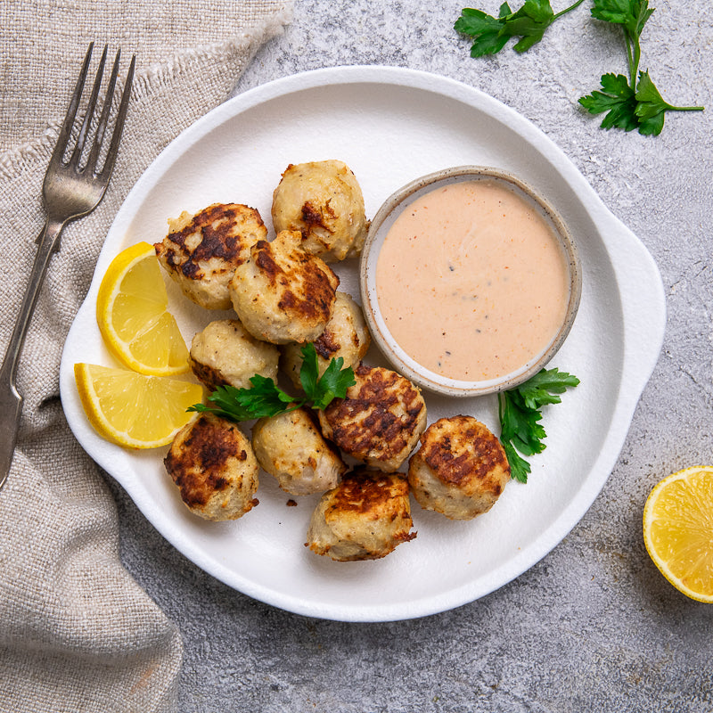 Plated popcorn chicken with dipping sauce, lemon wedges, and parsley on a gray surface.