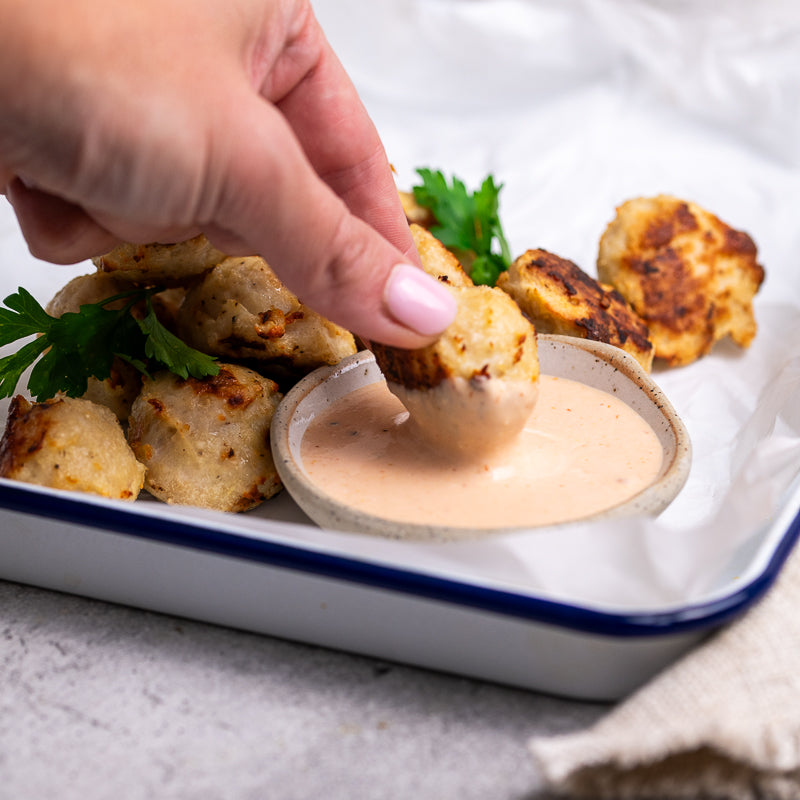 Person dipping a popcorn chicken bite into a creamy sauce with a plate of similar food in the background.