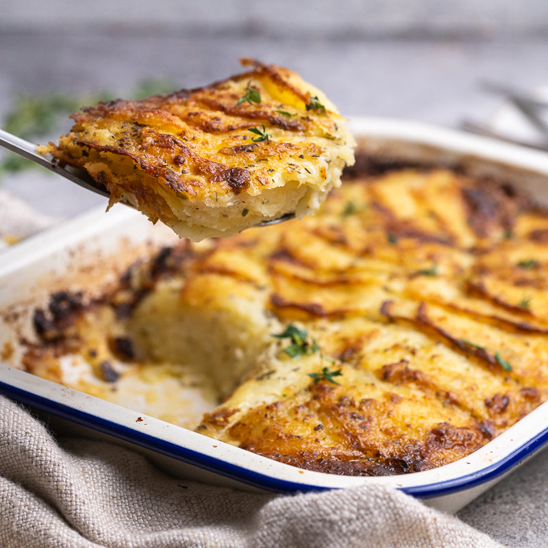 A close-up of a baked potato casserole or gratin being served on a spoon, with the remaining dish in a white baking dish on a rustic gray surface.